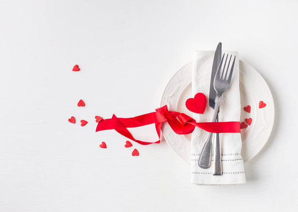 Valentines day (or wedding) meal background with red ribbon, hearts, fork, knife, white plate and napkin. Romantic holiday table setting. Beautiful background with blank. Restaurant concept. Flat lay - Zdjęcie, obraz