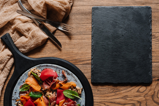Ensalada con mejillones, verduras de raíz y jamón, tenedor con cuchillo y pizarra en blanco sobre mesa de madera
 - Foto, Imagen