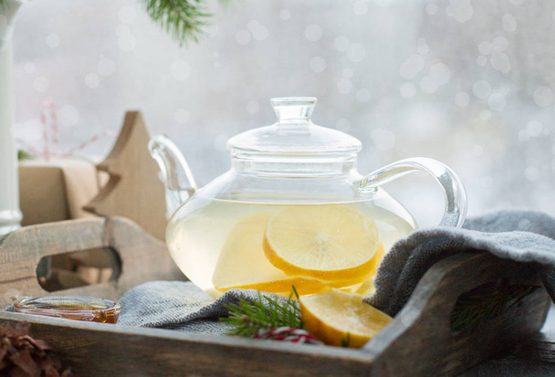 Hot green lemon ginger tea in a glass teapot on a wooden box in front of a window. Winter holidays concept. - Photo, Image