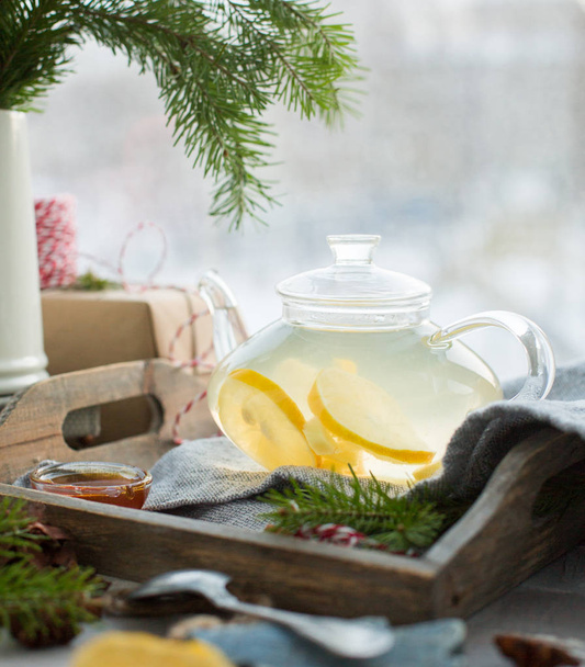 Heißer grüner Zitronen-Ingwer-Tee in einer gläsernen Teekanne auf einer Holzkiste vor einem Fenster. Winterferienkonzept. - Foto, Bild