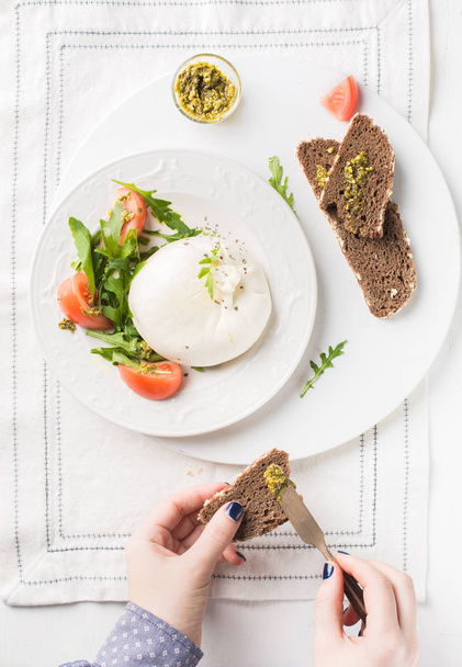 Sliced Italian cheese Burrata (mozzarella) with fresh arugula, tomatoes, olive oil, sauce pesto and whole grain bread toasts on a white plate and a wooden light background.Flat lay, top view - Photo, Image