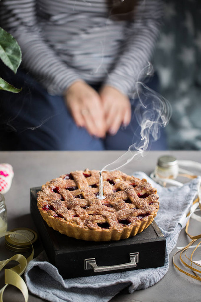 A birthday cherry cake with a burning one white candle. Atmospheric beautiful food birthday photo with home cherry pie, flowers, ribbons and decoration. - Photo, Image