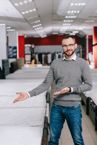 Porträt eines männlichen Kunden mit Brille, der auf arrangierte Matratzen im Möbelhaus zeigt - Foto, Bild