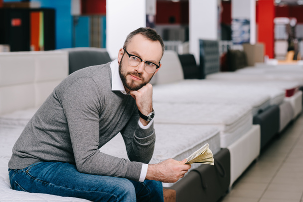 side view of pensive male shopper with dollar banknotes sitting on mattress in furniture store - Fotoğraf, Görsel