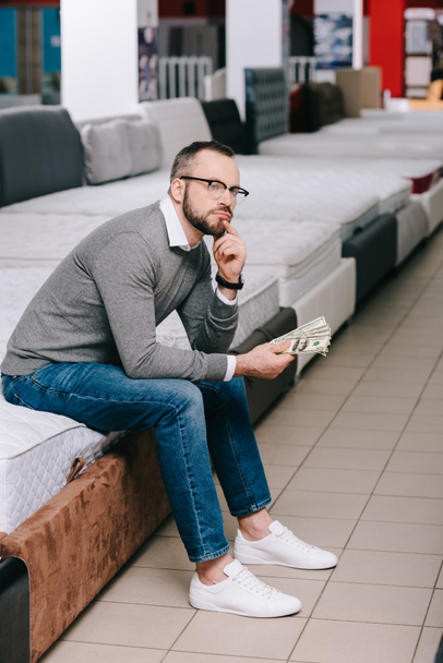 düşünceli erkek müşteri yatak mobilya mağazası içinde oturan Doları banknot ile yan görünüm - Fotoğraf, Görsel