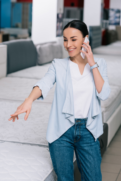 hymyilevä nainen puhuu älypuhelimella valitessaan patjaa huonekalukaupassa
 - Valokuva, kuva