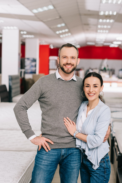 smiling couple looking at camera in furniture store with arranged mattresses - Photo, Image