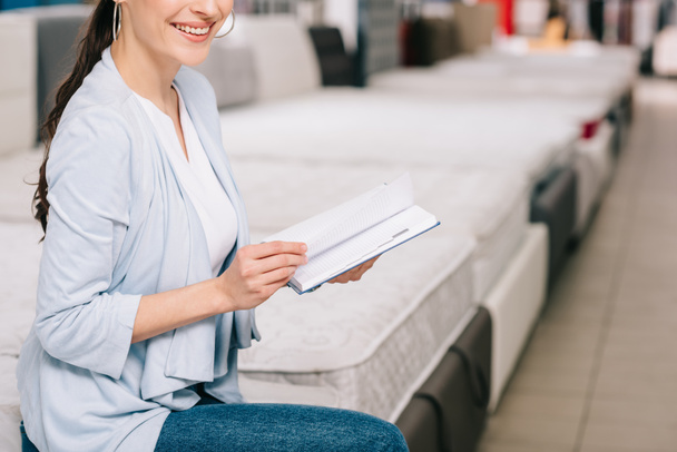 cropped shot of smiling female customer with notebook in furniture store with arranged mattresses - Photo, Image