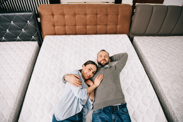 Εναέρια άποψη του χαμογελώντας ζευγάρι ξαπλωμένο στο κρεβάτι σε κατάστημα επίπλων με τοποθετημένα στρώματα - Φωτογραφία, εικόνα