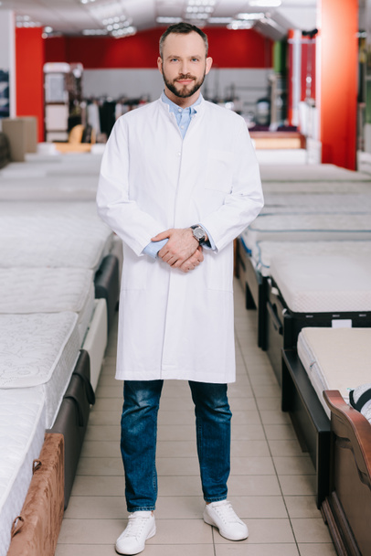 shop assistant in white coat standing in furniture shop with arranged mattresses - 写真・画像
