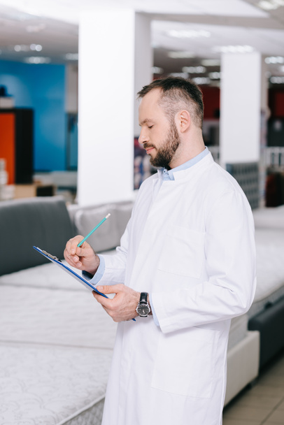 side view of shop assistant in white coat making notes in notepad in furniture shop with arranged mattresses - Photo, Image