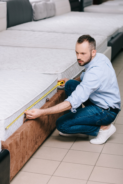 male shopper measuring orthopedic mattress with measure tape in furniture store - Foto, afbeelding