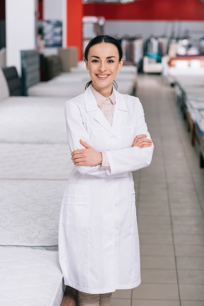 portrait of smiling shop assistant in white coat with arms crossed in furniture shop with arranged mattresses - Photo, Image