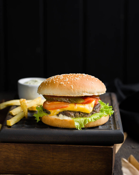 Delicious fresh tasty burger with beef, tomato, cheese and lettuce, french fries and beer served  on a wooden cutting board on dark background.Street fast food - Fotó, kép