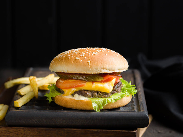 Heerlijke verse smakelijke hamburger met rundvlees, tomaat, kaas en sla, frietjes en bier geserveerd op een houten snijplank op donkere achtergrond. Straat fastfood - Foto, afbeelding