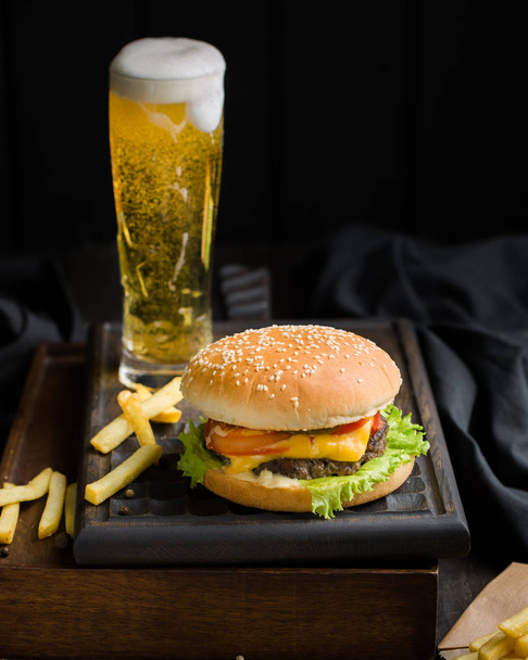 Delicious fresh tasty burger with beef, tomato, cheese and lettuce, french fries and beer served  on a wooden cutting board on dark background.Street fast food - Photo, Image