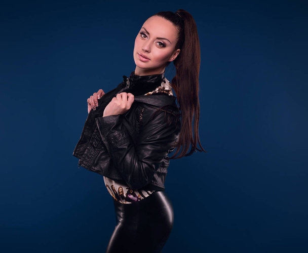 Portrait of young sexy girl with long hair in leather jacket on blue background in studio - Photo, Image