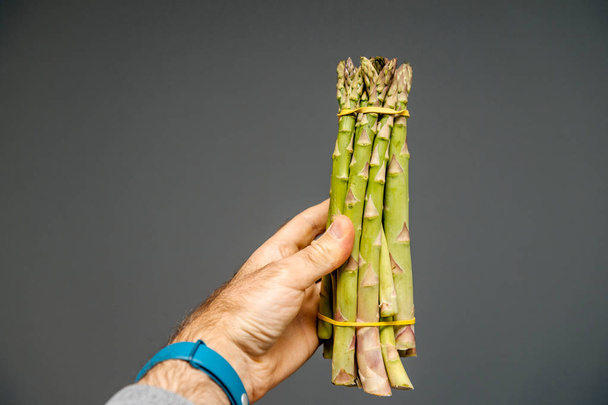 Male hand holding against gray background a bunch of fresh green asparagus - Zdjęcie, obraz