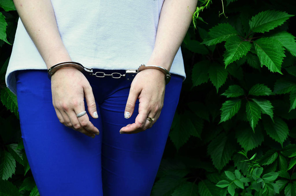 Fragment of a young criminal girl's body with hands in handcuffs against a green blossoming ivy leaves background. The concept of detaining an offender of a female criminal in a rural environment - Foto, Imagen