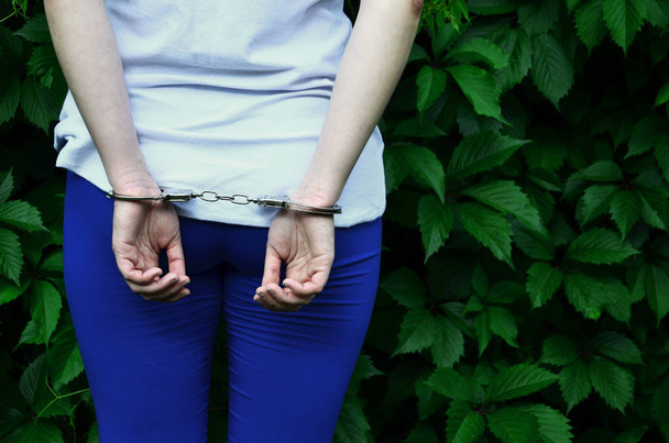 Fragment des Körpers eines jungen kriminellen Mädchens mit Händen in Handschellen vor einem grün blühenden Efeu hinterlässt Hintergrund. das Konzept der Inhaftierung eines Straftäters einer weiblichen Straftäterin im ländlichen Raum - Foto, Bild