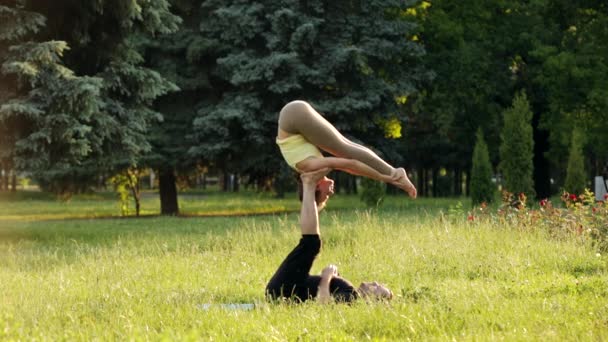 Hermosa pareja practicando acro yoga. Jóvenes instructores de yoga practican en un parque de la ciudad sobre hierba verde. Dos jóvenes exitosos realizan ejercicios de acro yoga. Hombre y una mujer aprenden yoga al atardecer
. - Imágenes, Vídeo
