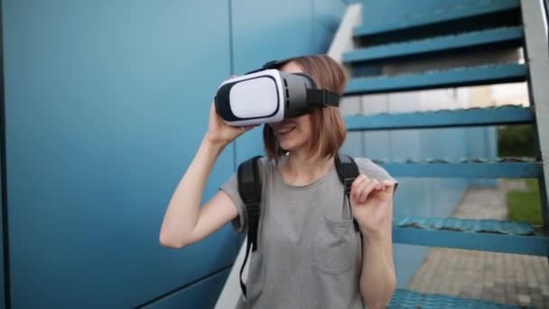 Будущее сейчас. Молодая женщина на лестнице играет в игру или смотрит кино в VR-очках. Молодая белая женщина прикасается к чему-то с помощью современных очков виртуальной реальности на синем фоне
. - Кадры, видео