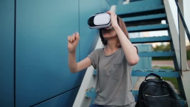Future is now. Beautiful young female on a stairs playing game in vr glasses. Young caucasian woman touch something using modern virtual reality glasses on a blue background. - Footage, Video