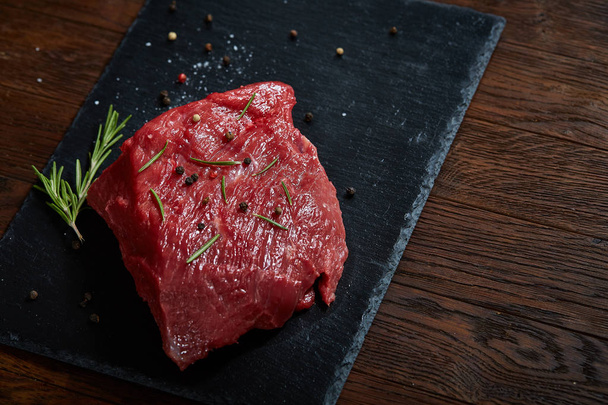 Raw meat beef steaks on black slate board with spices and rosemary over wooden background, copy space - Fotografie, Obrázek