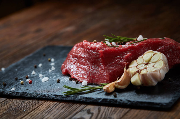 Raw meat beef steaks on black slate board with spices, garlic and rosemary over wooden background, selective focus - Fotografie, Obrázek
