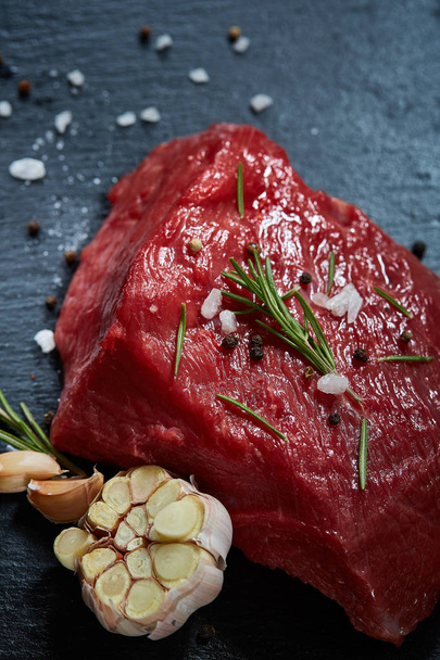 Raw meat beef steaks on black slate board with spices, garlic and rosemary over wooden background, selective focus - Foto, Imagem