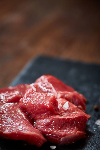Raw meat beef steaks on black slate board with spices and rosemary over wooden background, copy space - Zdjęcie, obraz
