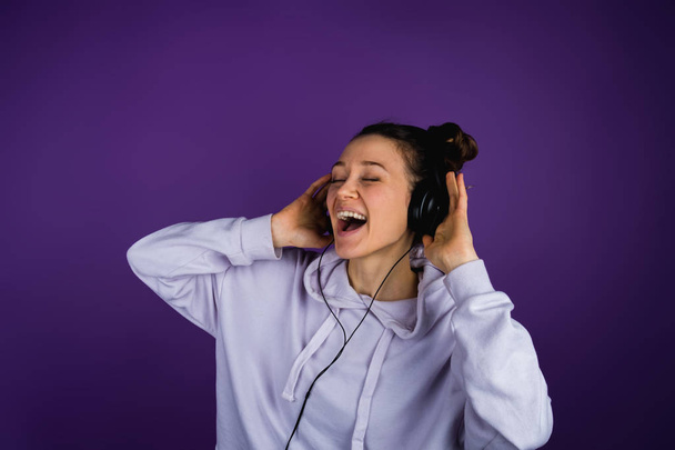 schönes junges Mädchen, das laut mit Kopfhörern in einem Sweatshirt auf lila Hintergrund singt - Foto, Bild