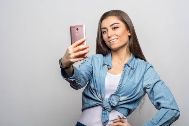 Portrait of a young smiling brunette woman in casual clothes making a selfie with her pink phone against white background. Lifestyle, people and technology concept - Φωτογραφία, εικόνα