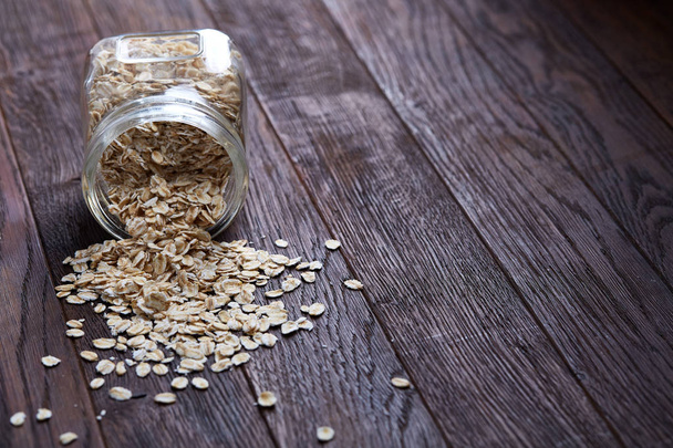 Overturned glass jar and spoon with raw oatmeal on vintage wooden background, close-up, selective focus. - Photo, image