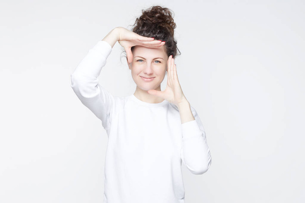 Indoor studio shot of  young attractive European female with curly hair bun smiling at camera and gesturing finger frame, while standing against isolated white wall background. Body language concept. - Photo, Image
