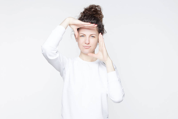Indoor studio portrait of beautiful young Caucasian woman with hair bun looking at camera and gesturing finger frame, while standing against isolated white wall background. Body language concept. - Photo, Image