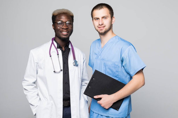 Best doctors. Two caucasian and afro american smiling doctors while standing against gray background - Foto, afbeelding
