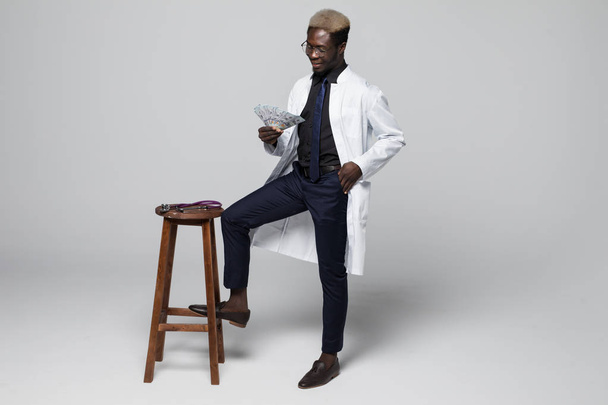 Portrait of handsome Afro American doctor in white coat looking at camera on chair holding dollars, isolated on graybackground - Photo, Image