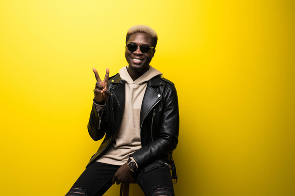 Portrét příležitostné afro americký mladík pózuje v sluneční brýle a ukázal vítězství znamení izolované žluté pozadí - Fotografie, Obrázek