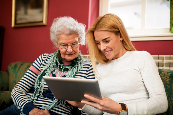 Kaksi sukupolvea käyttäen tablettia ja hymyillen yhdessä, tyttärentytär oppii isoäitiä käyttämään elektroniikkaa
 - Valokuva, kuva