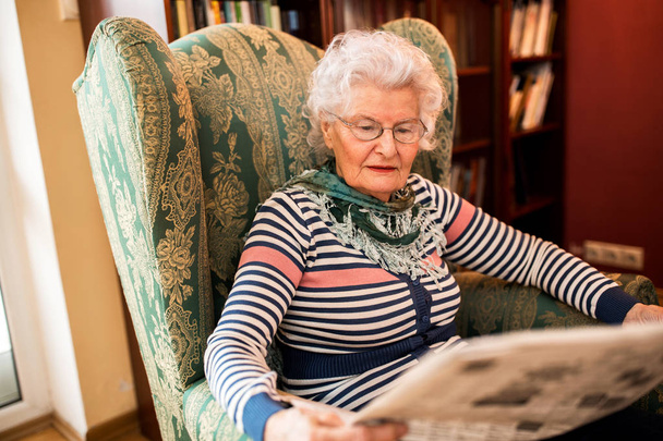 Senior old woman in pansion relaxing at home while reading a newspaper - Фото, изображение