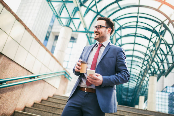Businessman holding mobile phone and cup of coffee outside of office buildings in the background.  - Foto, Bild