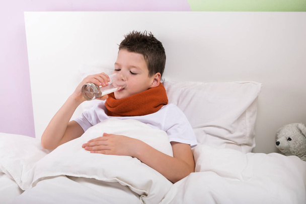 the boy drinks water from the glass from the temperature, lying on the bed - Foto, afbeelding