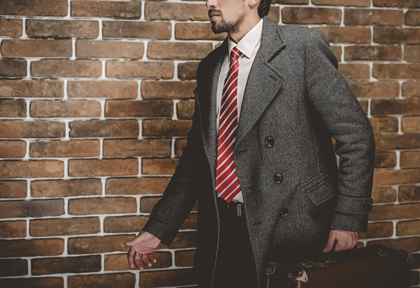 Portrét módní dobře oblečený muž s vousy pózuje venku koukal, jistý a soustředěný zralý muž v postavení kabát s kufrem na cihlové zdi pozadí, elegantní modelka. - Fotografie, Obrázek