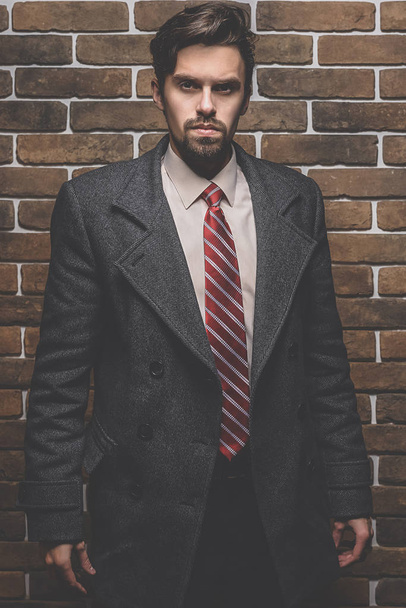 Portret van modieuze goed geklede man met baard poseren buitenshuis op zoek weg, zelfverzekerd en gerichte volwassen man in vacht staande op een bakstenen muur achtergrond, elegante fotomodel. - Foto, afbeelding
