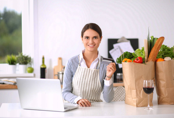 Femme souriante achats en ligne à l'aide d'un ordinateur et carte de crédit dans la cuisine - Photo, image
