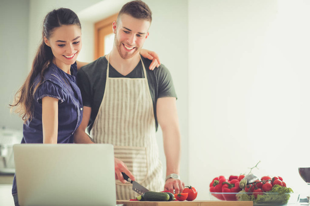 Young man cutting vegetables and woman standing with laptop in the kitchen - Photo, Image