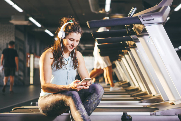 Beautiful young Caucasian girl sportswoman sitting, resting after training on treadmill against the backdrop of gym in sunny weather. Listens to music in sick white headphones,in hand holds the phone - Foto, imagen