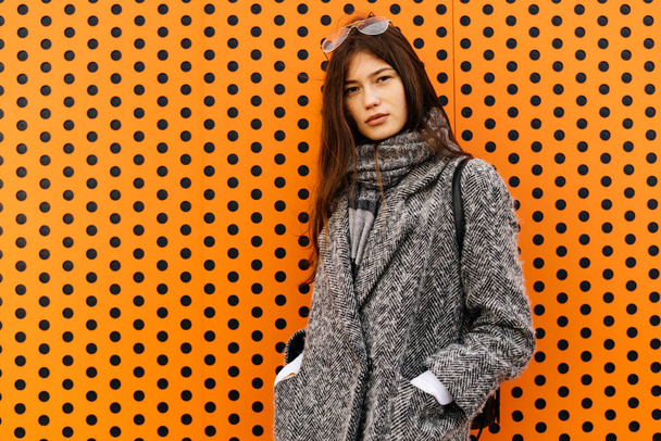 dark haired city girl in a trendy gray coat posing against an unusual orange wall - Фото, изображение