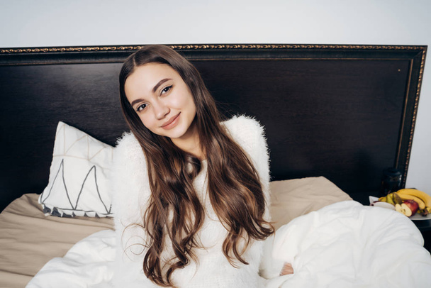 a cute young girl with long hair woke up early in the morning, sits in bed and a smiling new day - Photo, image